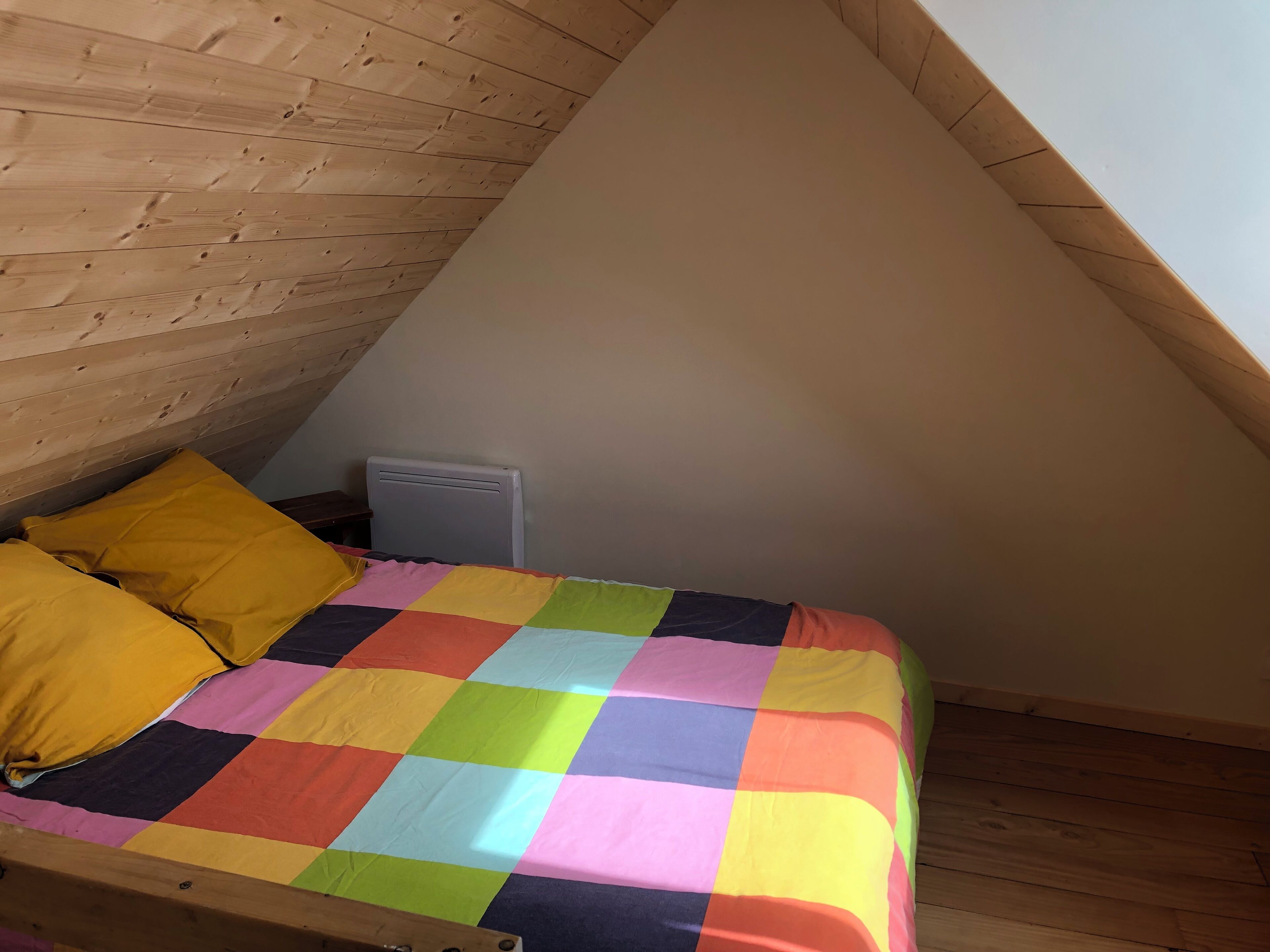 Una habitación pequeña y acogedora con techo inclinado, estructura de cama de madera, colcha de cuadros coloridos y un radiador.