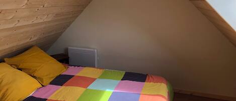 A small, cozy room with a sloped ceiling, a wooden bed frame, a colorful checkered bedspread, and a radiator.