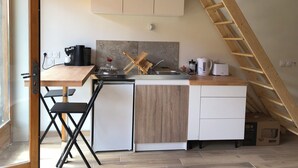 Eine kompakte Küche mit Holzboden, Spüle und kleinem Kühlschrank. Eine Holzleiter lehnt an der Wand, und eine Kiste mit der Aufschrift 'MOC25M' steht auf dem Boden.