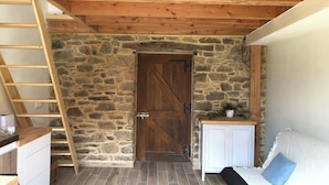 Ein rustikaler Raum mit Steinwand, Holzbalkendecke, Holztür, weißem Schrank und einem Bett mit blauem Kissen. Eine Holzleiter ist ebenfalls zu sehen.