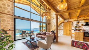 A spacious living room with a large window offering a view of the sea, a cozy seating area, and a wooden staircase leading to an upper level. - Rustic Villa Perfect for Couples (Bezirgan)