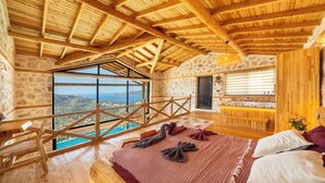 A high-ceilinged room with a wooden ceiling and stone walls, a large window offering a view of the landscape, a wooden bench, and a bed with pillows and a blanket. - Rustic Villa Perfect for Couples (Bezirgan)
