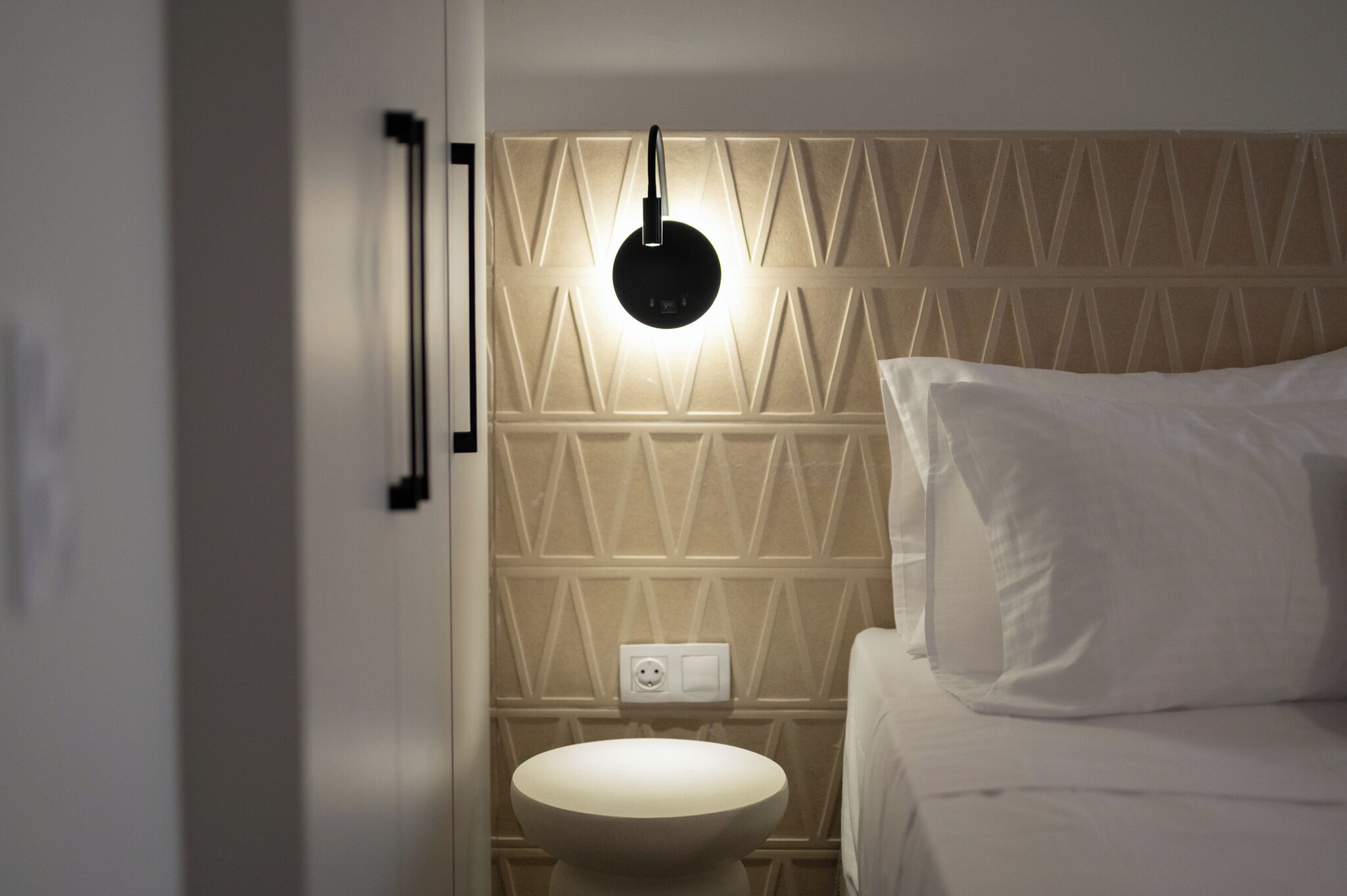 A modern bathroom with a geometric-patterned wall, a round sink, and a bedside table with a white pillow.
