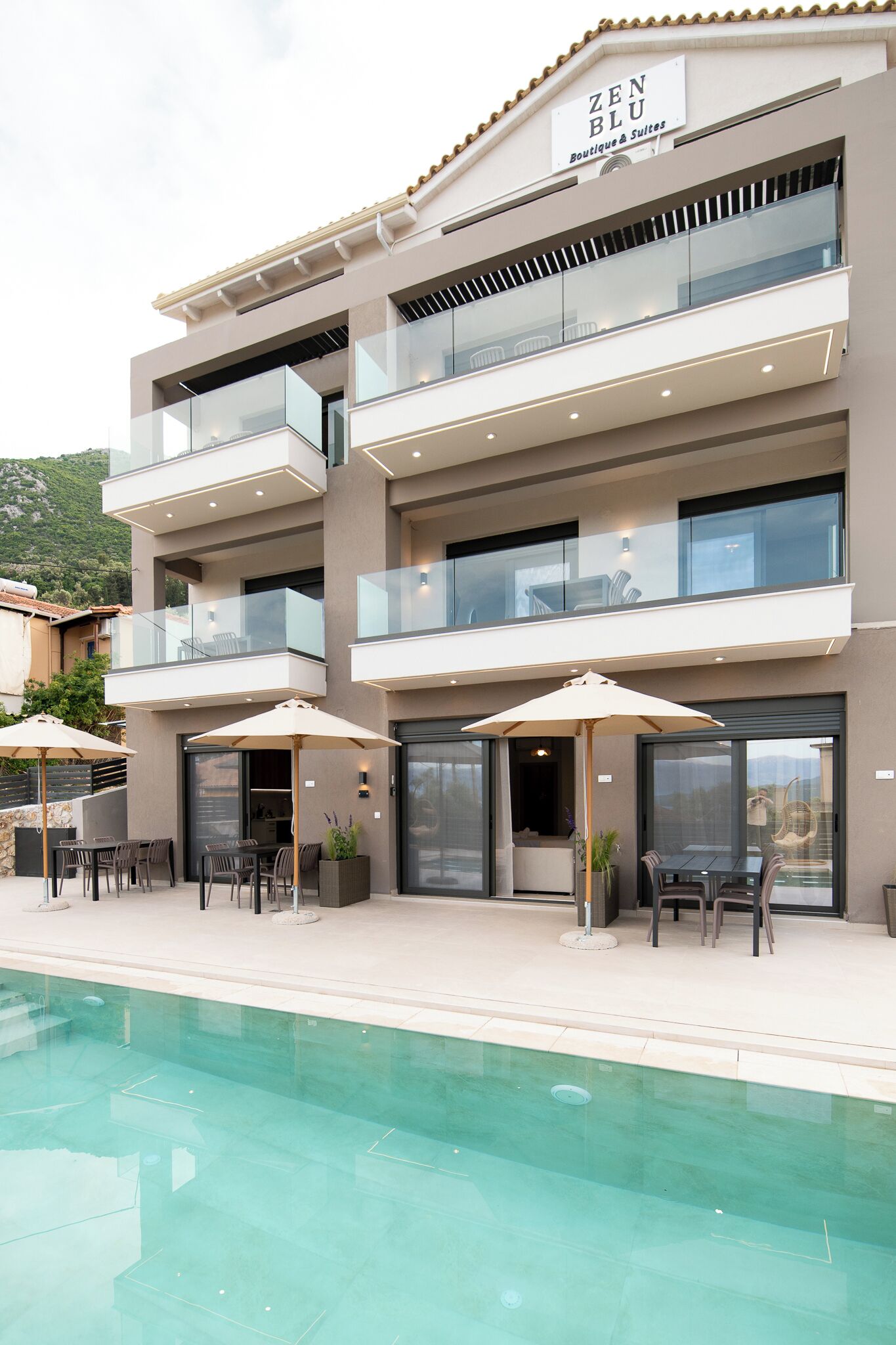 Un immeuble à plusieurs étages, sur lequel on peut lire l’inscription « ZEN BLU Boutique & Suites ». Il y a des espaces de détente en plein air avec des parasols et une piscine à l’avant.