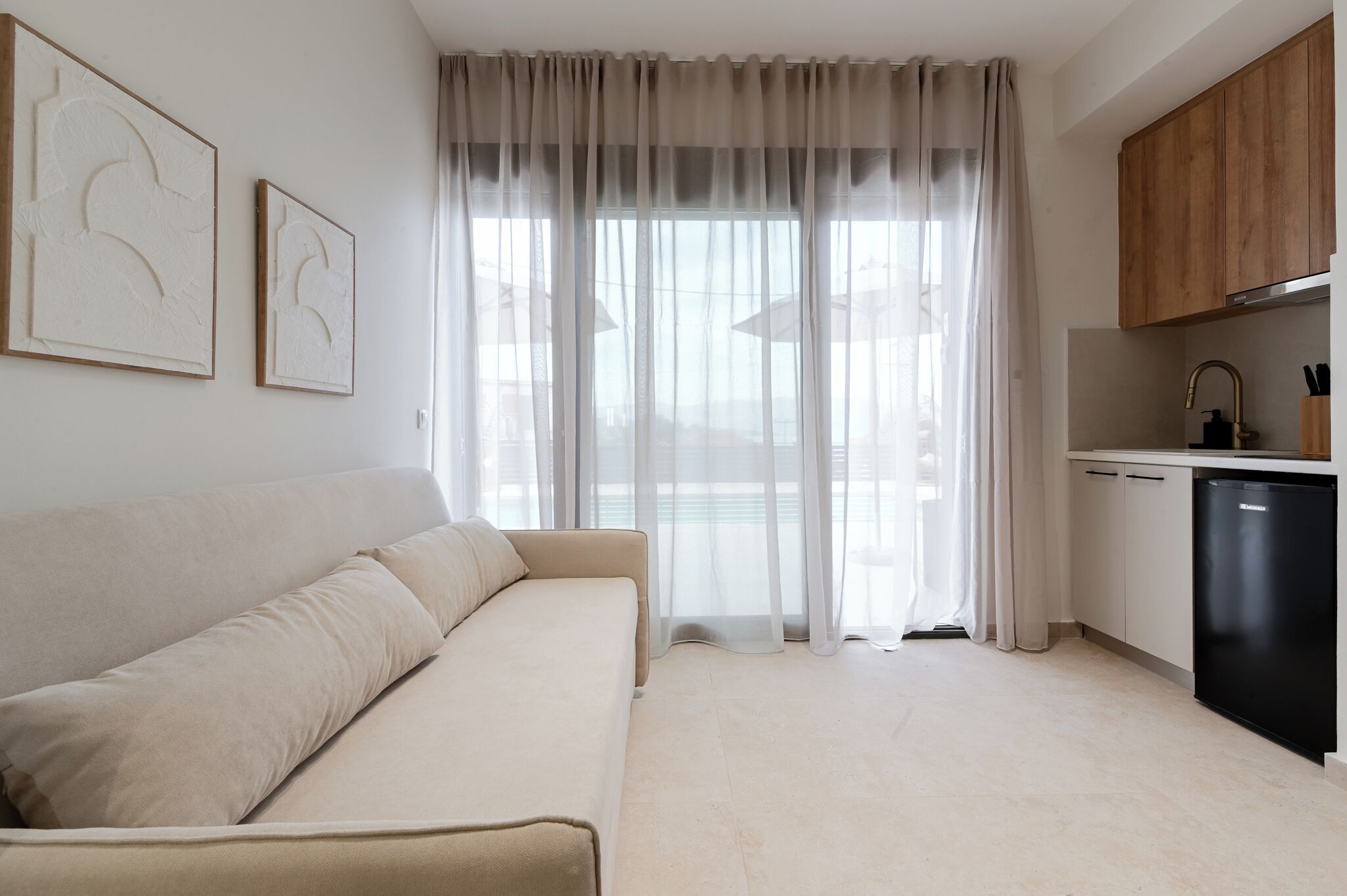 Una sala moderna con un sofá color crema, una cocina con armarios de madera y cortinas de tela ligera.