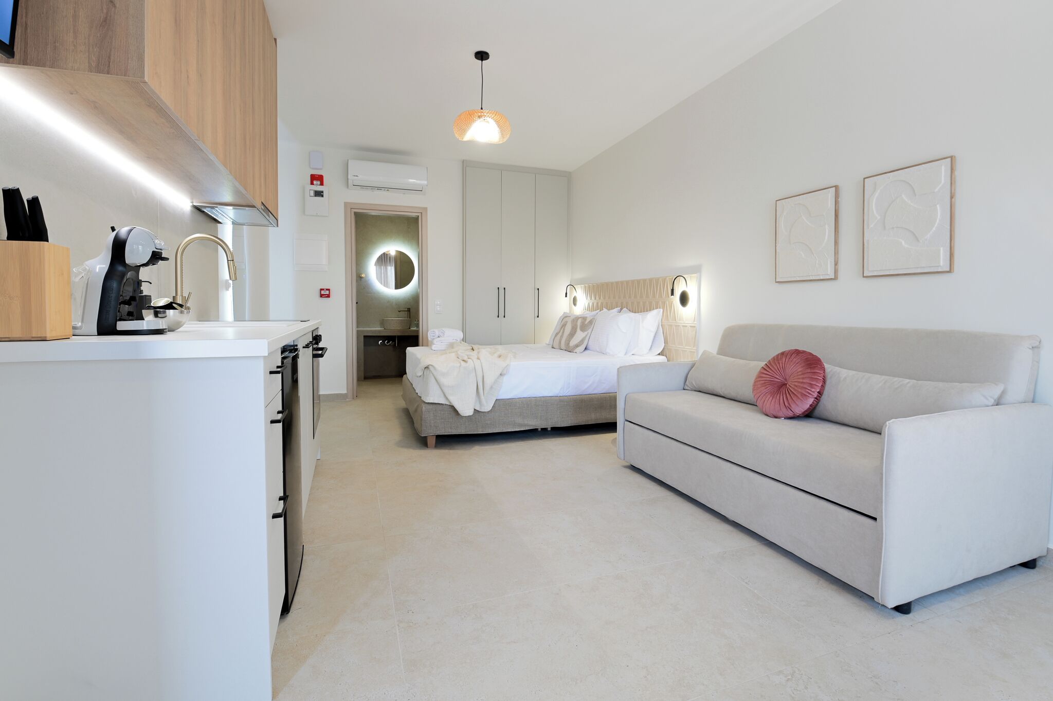 A modern hotel room with a kitchenette, a bed with a headboard, a sofa, and a pendant light.