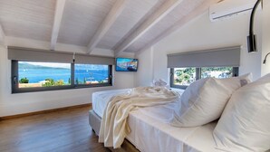 A bedroom with a bed, two windows with views of the sea, a mounted TV, and an air conditioning unit. - ZEN BLU Suites (Nikiana)