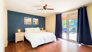Une chambre à coucher comprenant un lit, deux tables de chevet, un ventilateur de plafond et une grande fenêtre avec des rideaux.