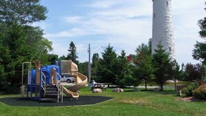 Une aire de jeux avec un toboggan et des structures d’escalade, un phare en arrière-plan et une table de pique-nique.