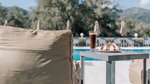 A poolside table with a drink and sandwiches, surrounded by lounge chairs and umbrellas, with a pool and trees in the background. - Pelithea Aparthotel (Corfu)