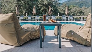 Uma área de estar à beira da piscina com duas cadeiras bege acolchoadas, uma mesa com bebida e pão, e guarda-sóis.