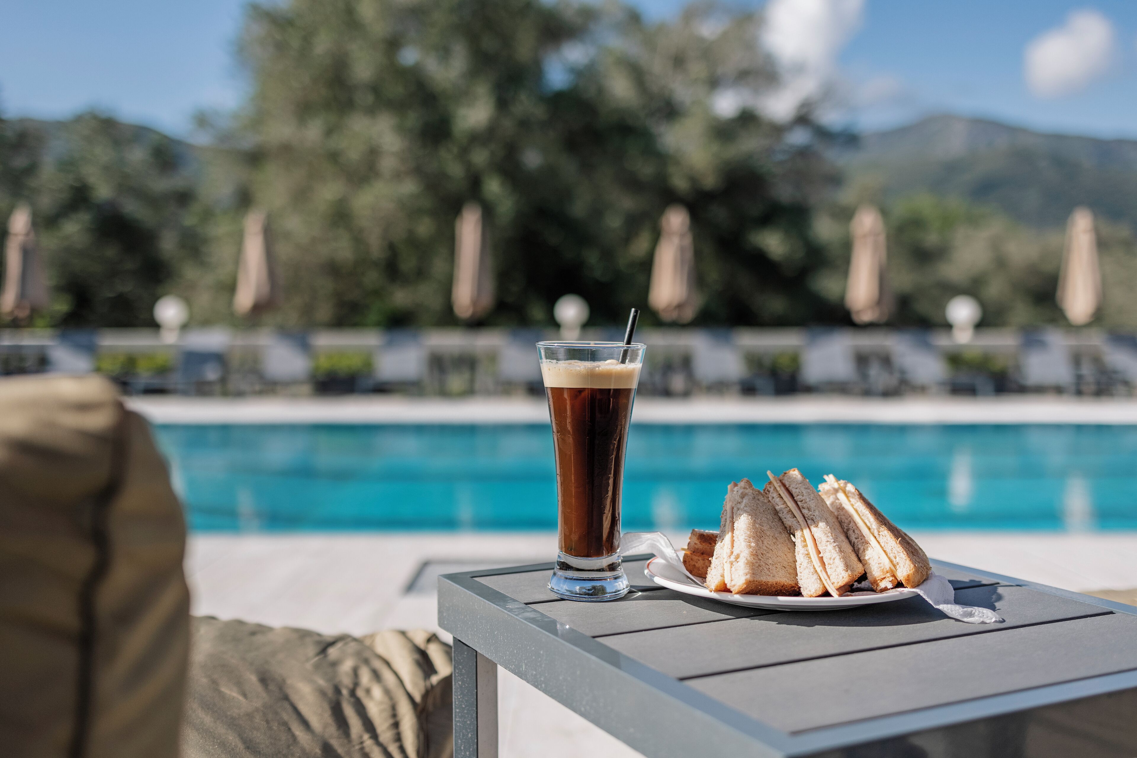 Un verre de café glacé et des sandwichs sur une table près d’une piscine, avec des chaises longues et des parasols en arrière-plan.