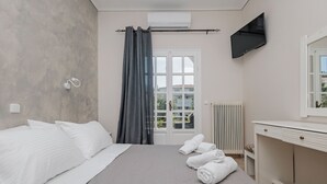A bedroom with a bed, a TV, a window with curtains, a radiator, and a desk with drawers. - Pelithea Aparthotel (Corfu)