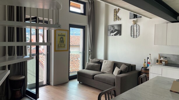 A modern living room with a grey sofa, a dining table, and a kitchen area. There is a staircase with a view of the outside, a door with a poster, and a wall with a large letter 'K'. - Open-plan loft overlooking Lake Como with private parking space (Malgrate)