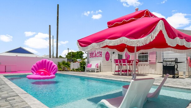A swimming pool with pink and white chairs, a pink flamingo float, and a beach-themed building with a red umbrella. - Beach House - Pink Prerogative by PHG (Panama City Beach)