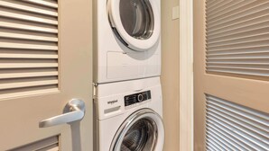 Une machine à laver et un sèche-linge superposés dans une pièce aux murs de couleur beige et aux fenêtres équipées de stores.
