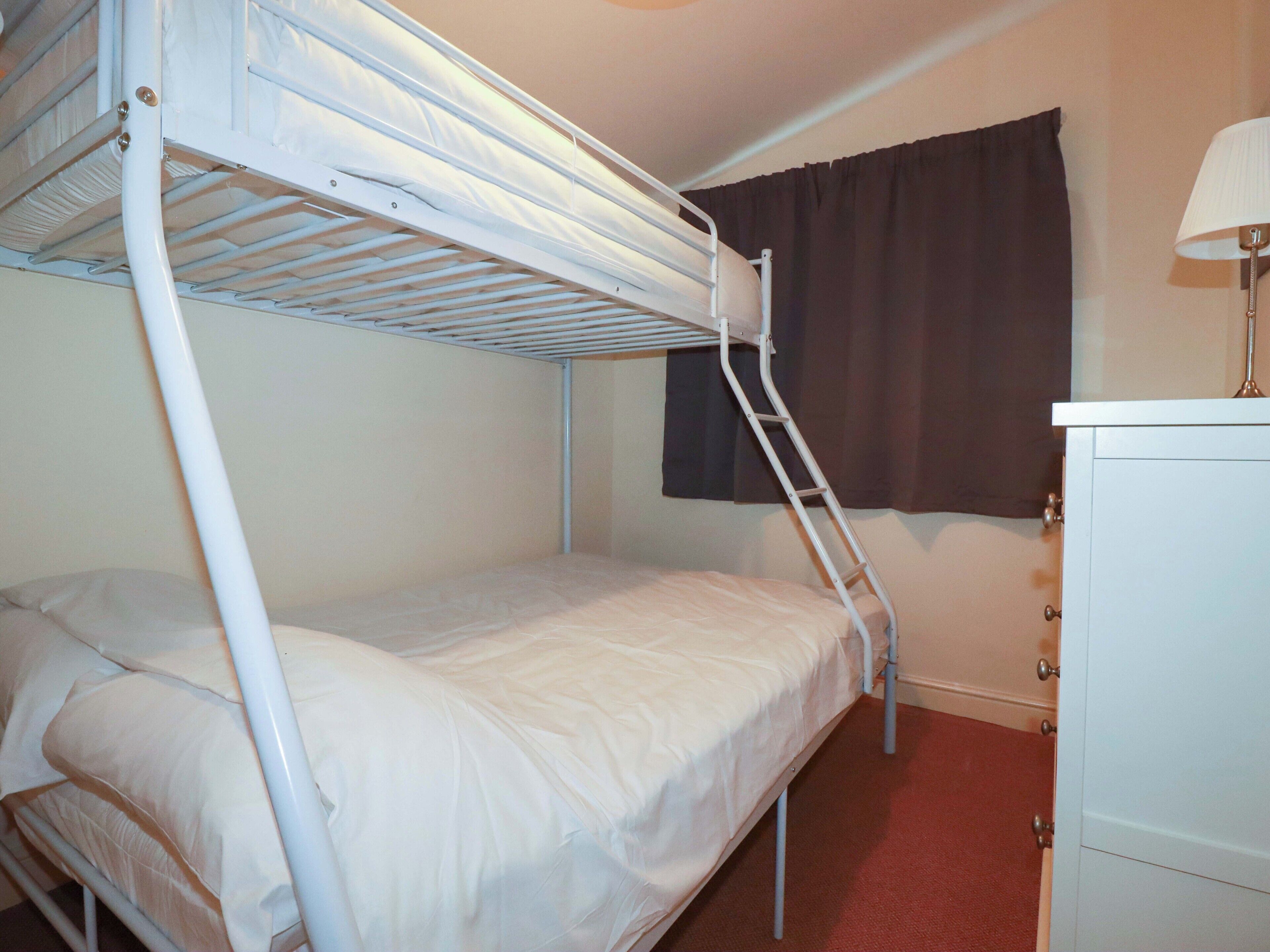 Una cama nido con una escalera, un pequeño aparador y una ventana con cortinas.