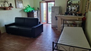 Ein Zimmer mit einer schwarzen Couch, einem Bett mit Metallgestell, einem Kühlschrank, einem kleinen Tisch mit Spiegel und einem Regal mit verschiedenen Gegenständen.