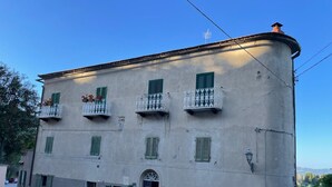 Ein zweistöckiges Gebäude mit grünen Fensterläden, einem Balkon mit weißen Geländern und einem kleinen Gartenbereich mit Topfpflanzen.