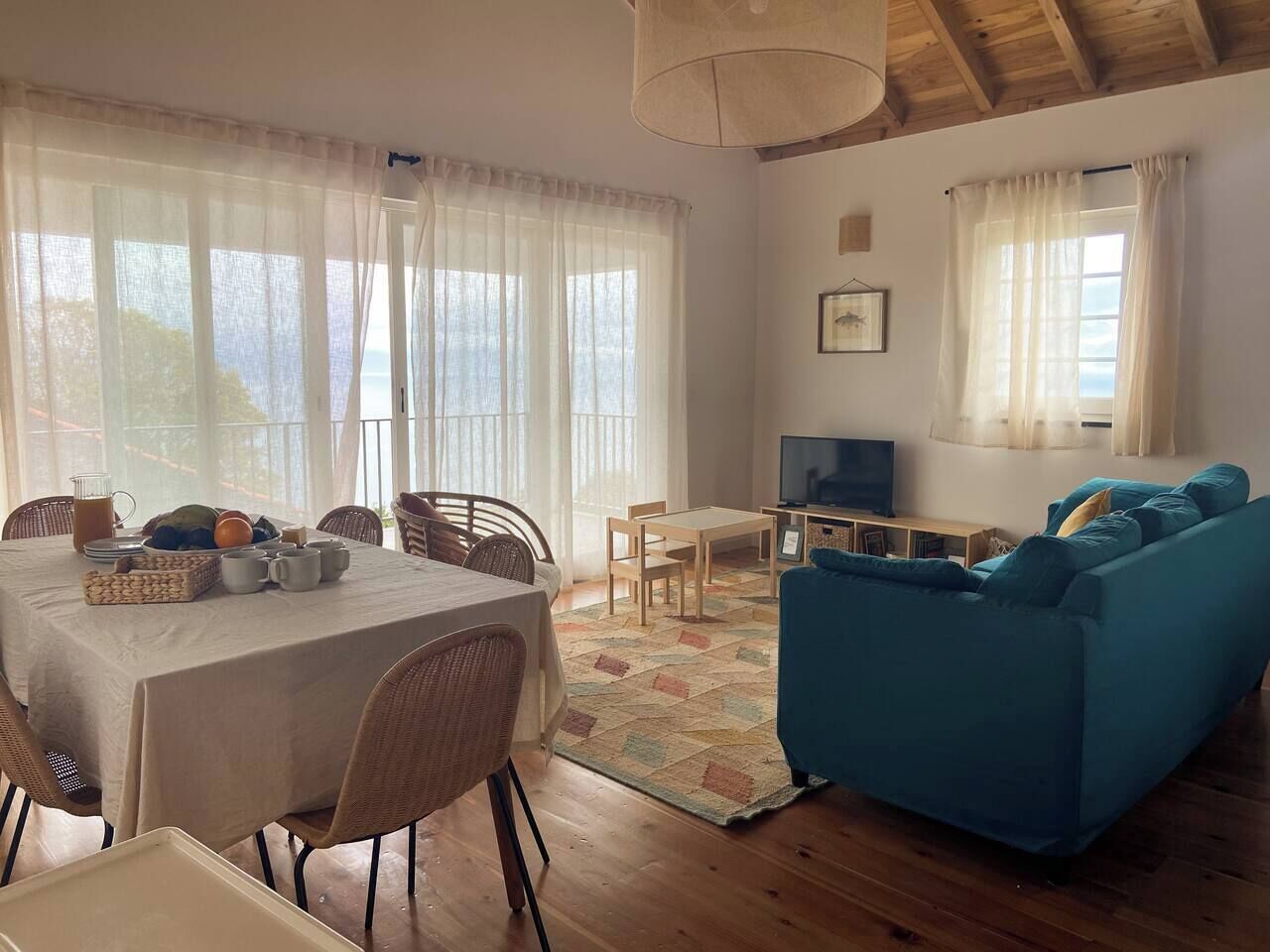 A cozy living room with a dining area, wooden flooring, and large windows with sheer curtains.