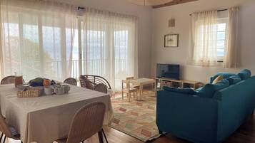 A cozy living room with a dining area, wooden flooring, and large windows with sheer curtains.