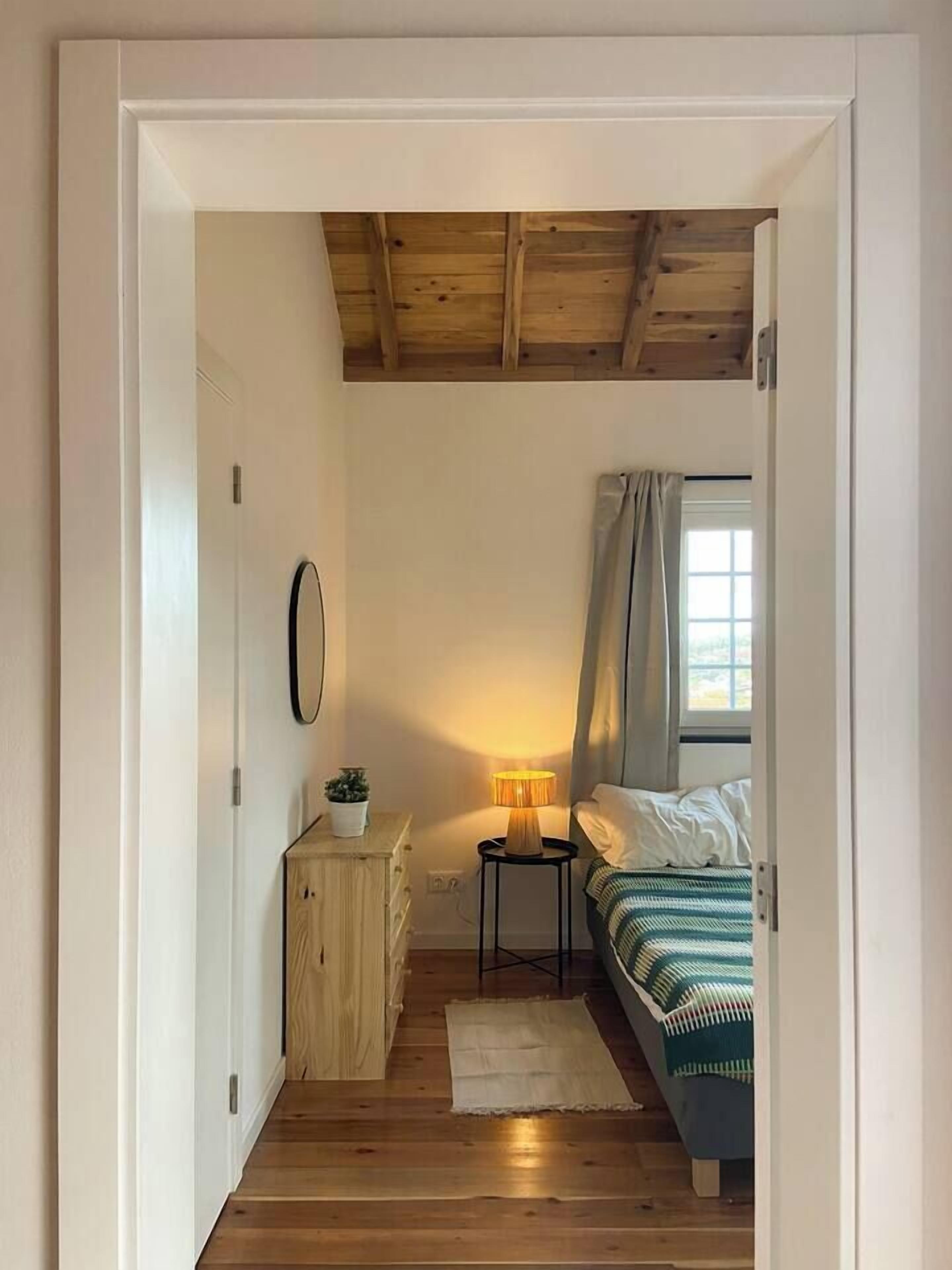 A bedroom with a bed, a bedside table with a lamp, a small cabinet, and a window with curtains.