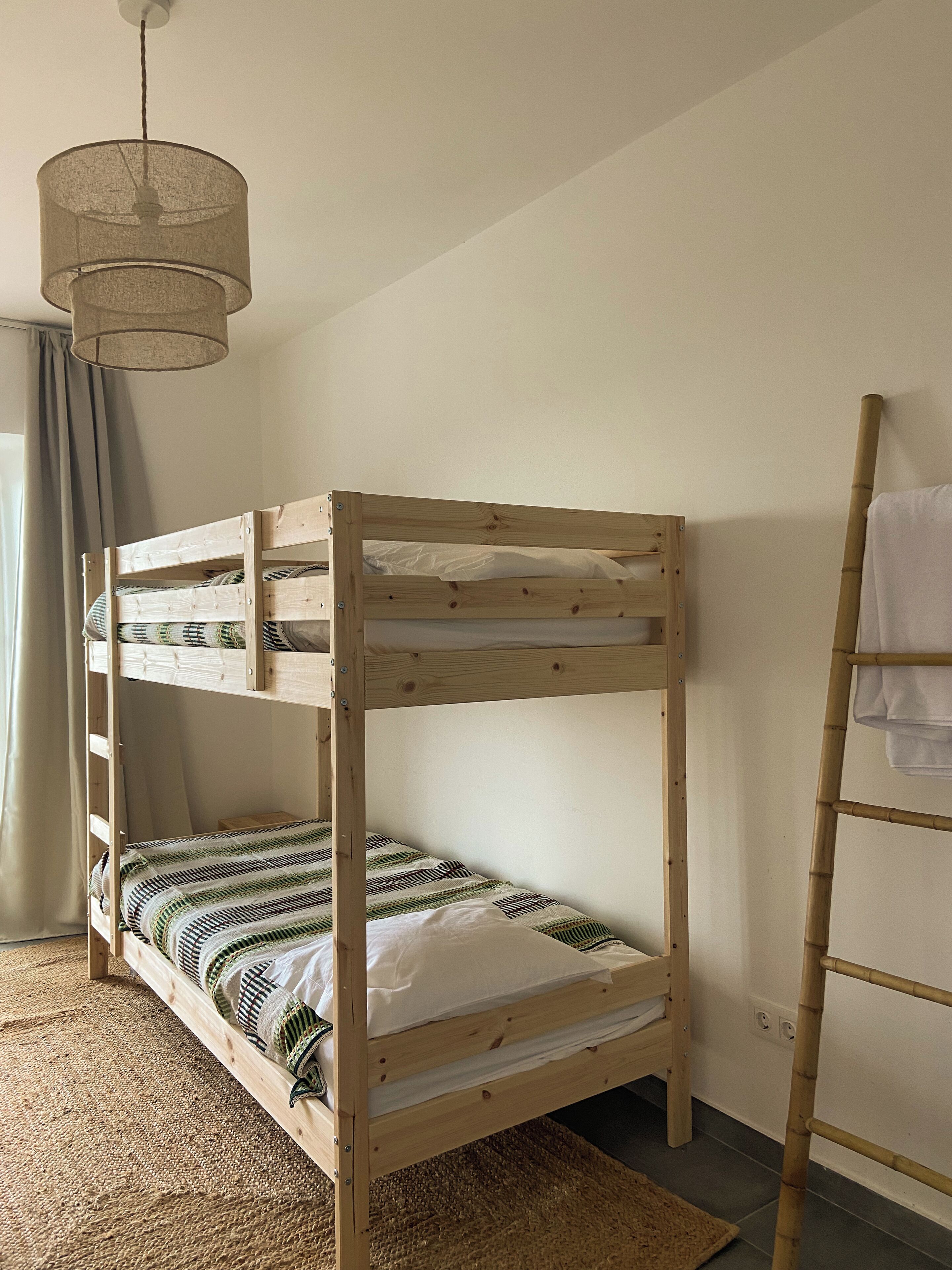 A bunk bed with a wooden frame, a woven headboard, and a woven footboard. There is a ladder on the right side and a hanging light fixture above the bed.
