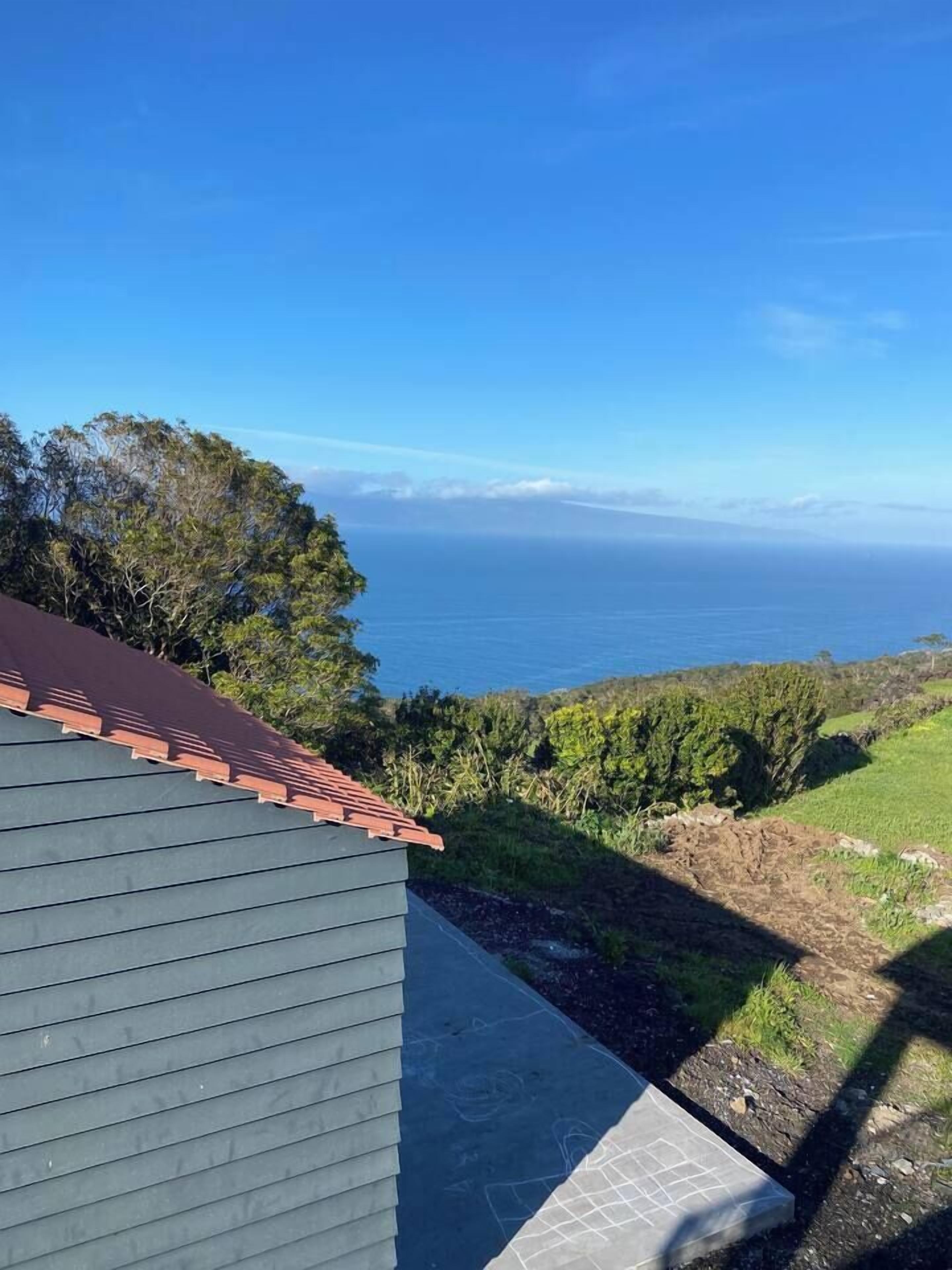 A house with a red roof and grey siding, overlooking a coastal view with the ocean and greenery.