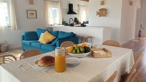Un coin repas avec une table mise pour le petit-déjeuner, un canapé bleu en arrière-plan et une cuisine visible à travers une porte ouverte.