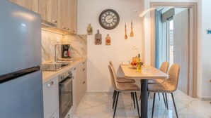 A modern kitchen with a built-in refrigerator, oven, and sink. There is a dining area with a table and chairs, and a wall-mounted clock. - Villa Leandros Premium Suites (Agia Varvara)