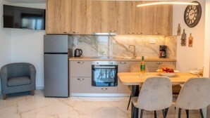 A compact kitchen with wooden cabinets, a built-in oven, a marble backsplash, and a dining area with a table and chairs. - Villa Leandros Premium Suites (Agia Varvara)