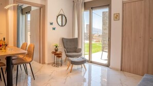 A modern room with a dining table, chairs, a grey armchair, a small side table with a vase, a mirror, and a view of the outdoors through a sliding door. - Villa Leandros Premium Suites (Agia Varvara)