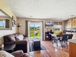 A living room with a sofa, a dining table with chairs, a kitchen area with cabinets, and a large window with a view of greenery.