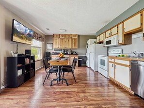 A modern kitchen with wooden cabinets, a dining table with chairs, a TV mounted on the wall, and a coffee bar.