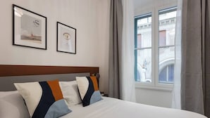 A hotel room with a large bed, two framed pictures on the wall, and a window with a view of buildings. - Chic Trieste Gem (Trieste)