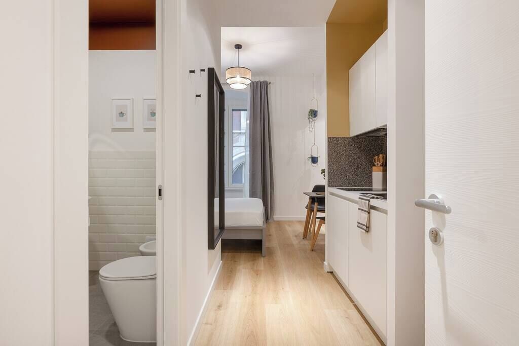 Un pasillo estrecho con un inodoro blanco, una cama, una mesita pequeña y una kitchenette con encimera y armarios.