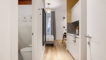 Un pasillo estrecho con un inodoro blanco, una cama, una mesita pequeña y una kitchenette con encimera y armarios.