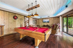 Una mesa de billar en una sala con paredes de madera, techo con vigas de madera y un ventanal grande.