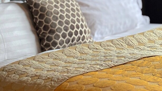 Una cama con cabecera estampada, almohadas blancas y colcha con motivos dorados.