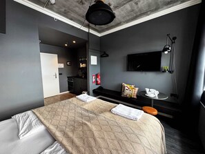 Quarto de hotel moderno com uma cama grande, um pequeno espaço de estar, televisão e cozinha compacta.