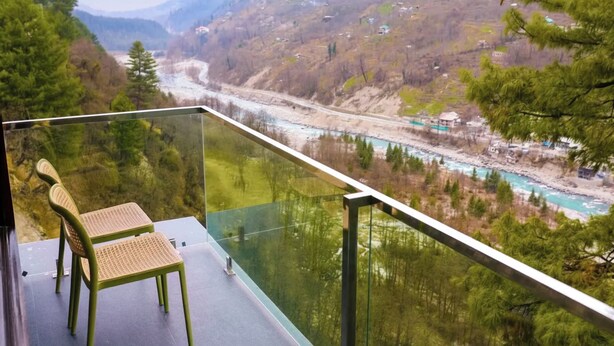 Un balcon avec deux fauteuils en osier donnant sur une vallée traversée par une rivière, avec des maisons et des arbres.