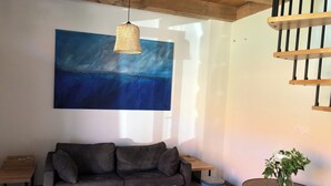 Un soggiorno con un divano grigio, un tavolino da caffè bianco, un quadro astratto blu, un lampadario in vimini appeso, un tavolino laterale in legno, un piccolo tavolino rotondo con un vaso e una scala con ringhiere in legno.