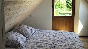 Camera da letto con soffitto spiovente, letto con copriletto decorato, finestra vista su vegetazione e porta in legno.