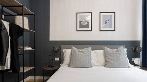 A hotel room with a bed, two pillows, a shelf with clothes, and two framed pictures on the wall. - Trieste Tranquil Tiny Haven (Trieste)