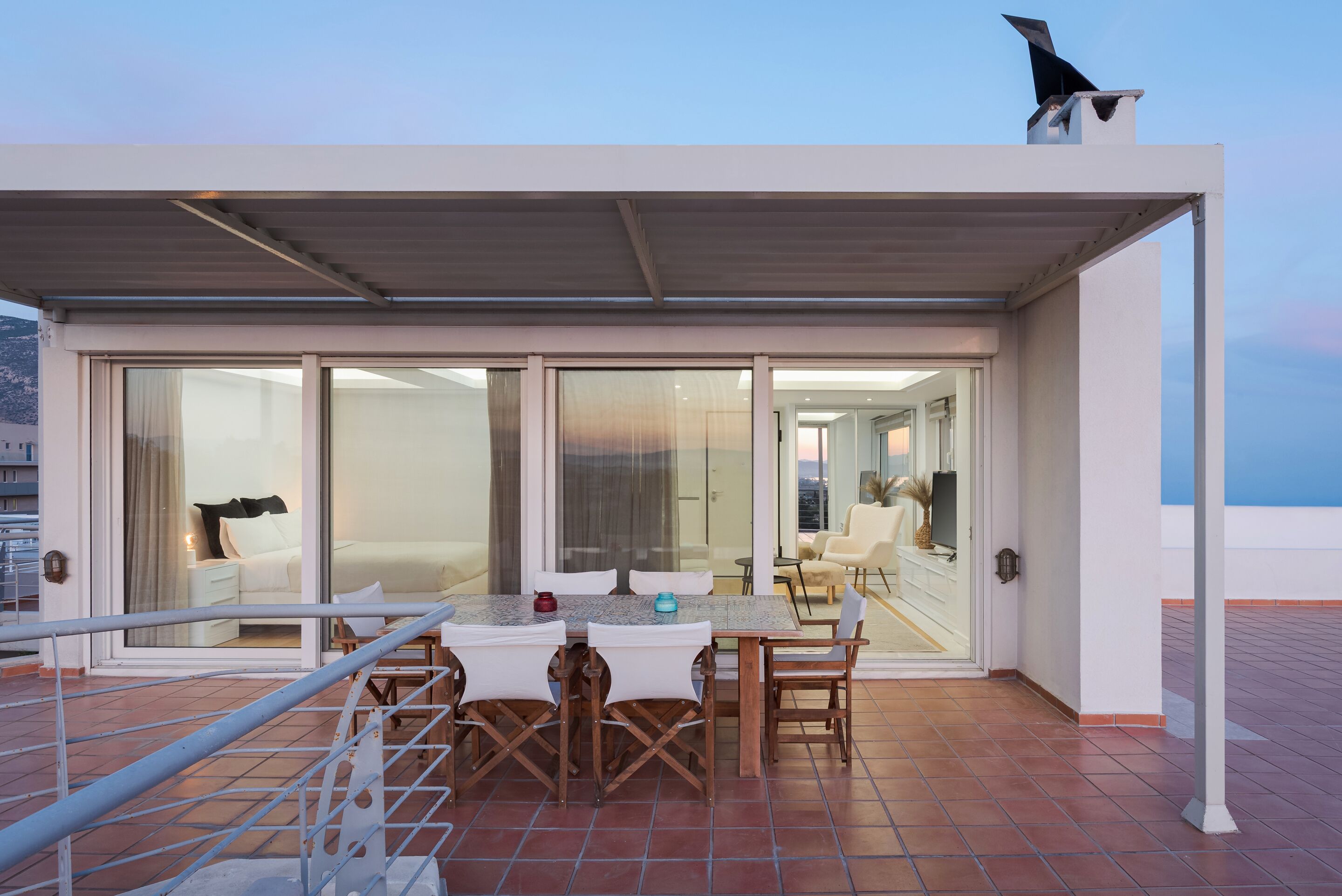 A covered outdoor seating area with a table and chairs, a railing, and a view of the interior through glass doors.