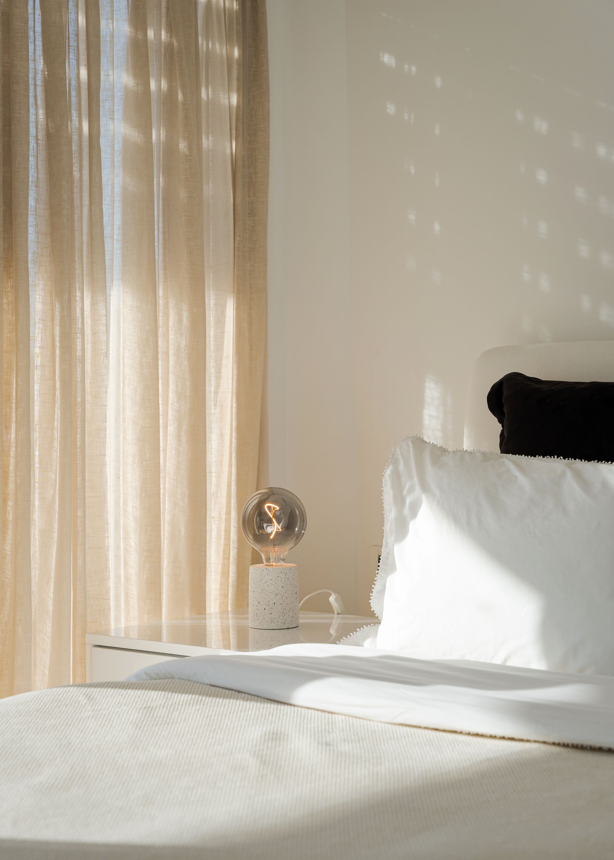 Ein ordentlich bezogenes Bett mit weißer Tagesdecke und Kissen, ein kleiner Tisch mit einer Lampe und ein Fenster mit durchsichtigen Vorhängen, das natürliches Licht hereinlässt.