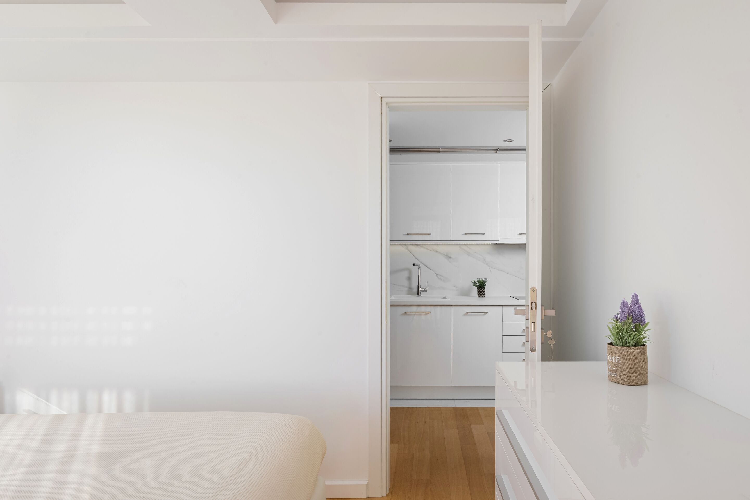 A modern bedroom with a bed, a small bedside table, and a nightstand. There is a doorway leading to a kitchen with white cabinets and a marble countertop.