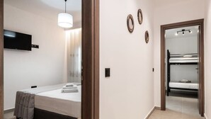 Une chambre comprenant un lit, une table de chevet, un téléviseur fixé au mur et une porte donnant sur un espace avec des lits superposés.