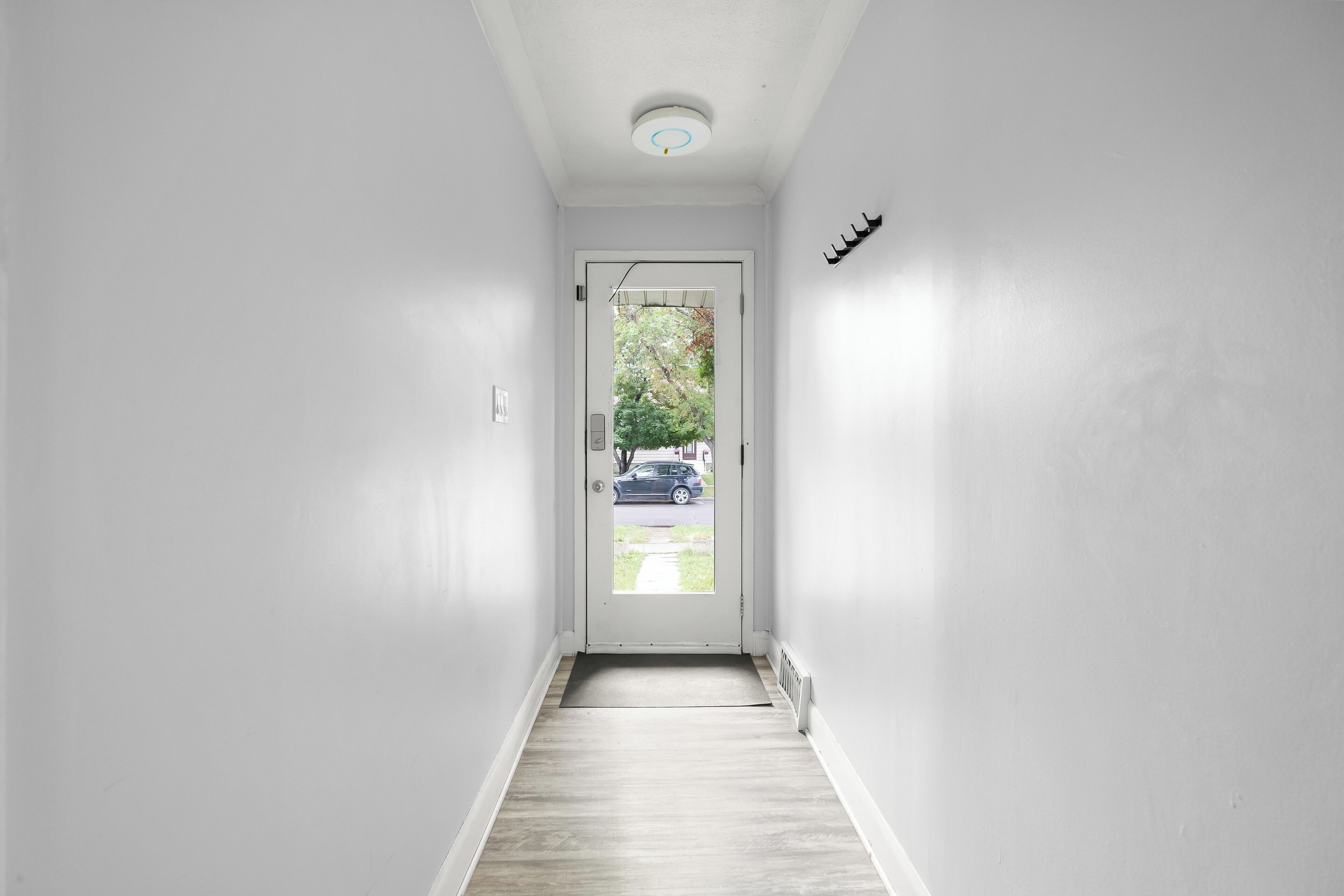 A narrow hallway with a white door at the end, a small step leading outside, and a visible tree and parked car through the door.