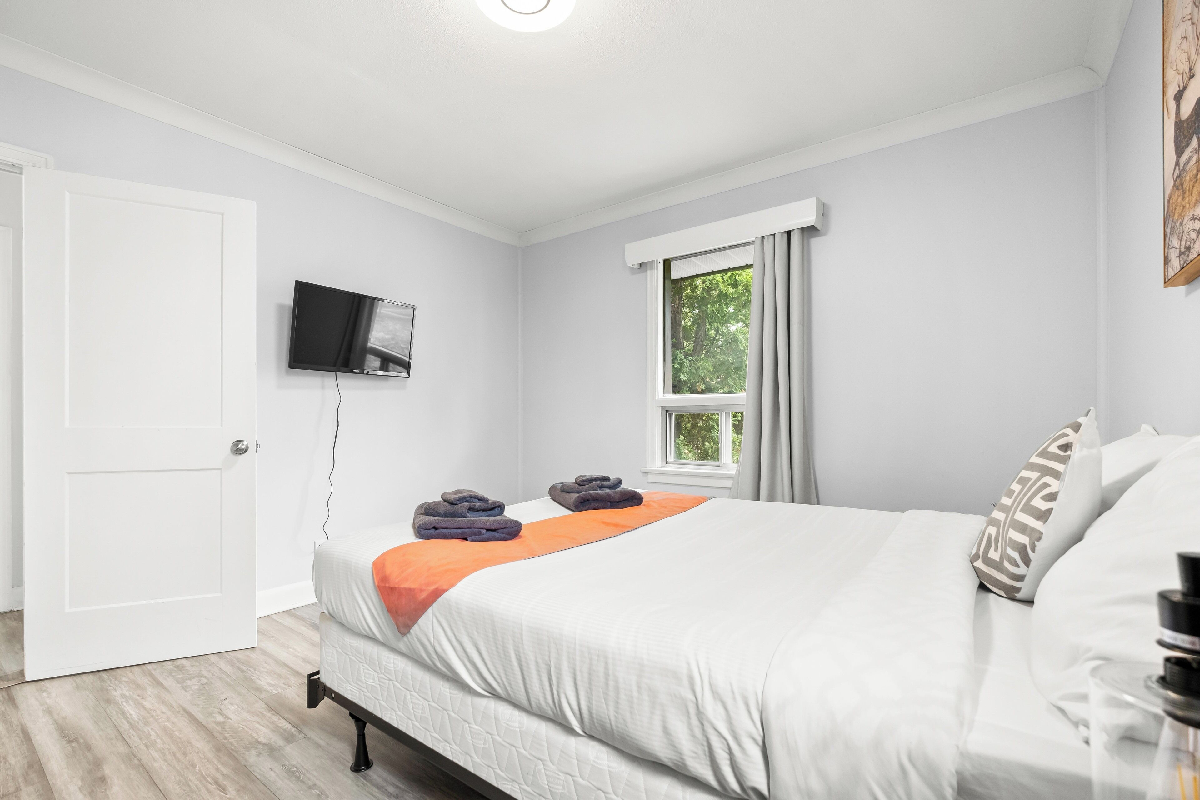 Ein ordentlich eingerichtetes Schlafzimmer mit einem Bett, einem an der Wand montierten Fernseher, einem Fenster mit Vorhängen und einer Tür.